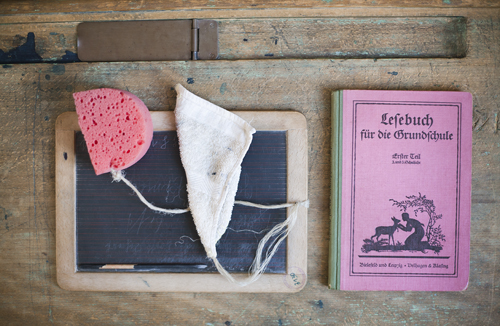 Auf einer Schulbank liegt eine Schiefertafel mit Lappen, Schwamm und Griffel. Rechts daneben liegt ein pinkes Lesebuch für die Grundschule.