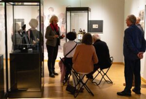 Auf diesem Foto ist eine Museumsführung dargestellt. Auf der linken Seite steht eine erzählende Frau, um sie herum sitzen Menschen in Stühlen und lauschen ihr gespannt.